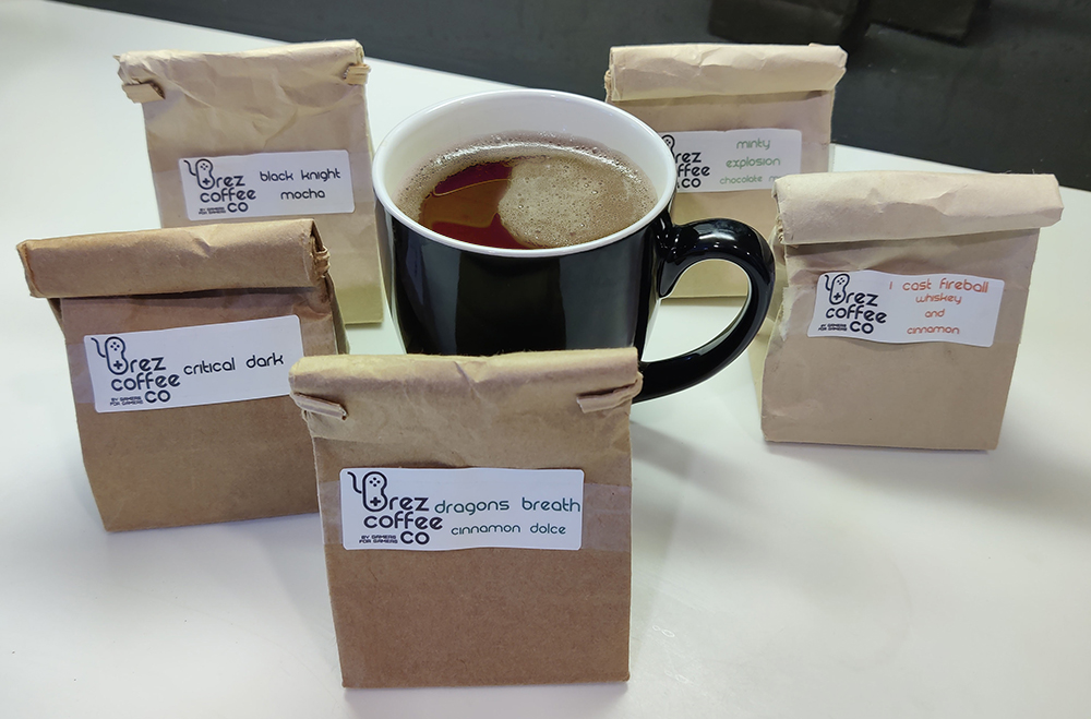 Black coffee mug filled with coffee. Surrounded by 5 small brown bags of sample coffee on a white countertop